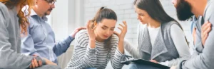 A distressed woman in a striped shirt, sitting with her head in her hands, surrounded by a supportive group during a drug rehab intervention discussion.
