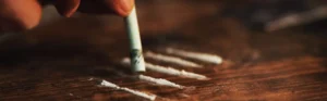 A person's hand holding a rolled dollar bill is about to snort coke lines on a wooden table, representing substance abuse before entering drug rehab treatment.
