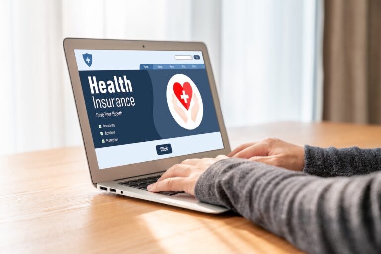 A person in a gray sweater is typing on a laptop displaying a "Health Insurance" website with a heart icon, checking coverage options for medical care and treatment.