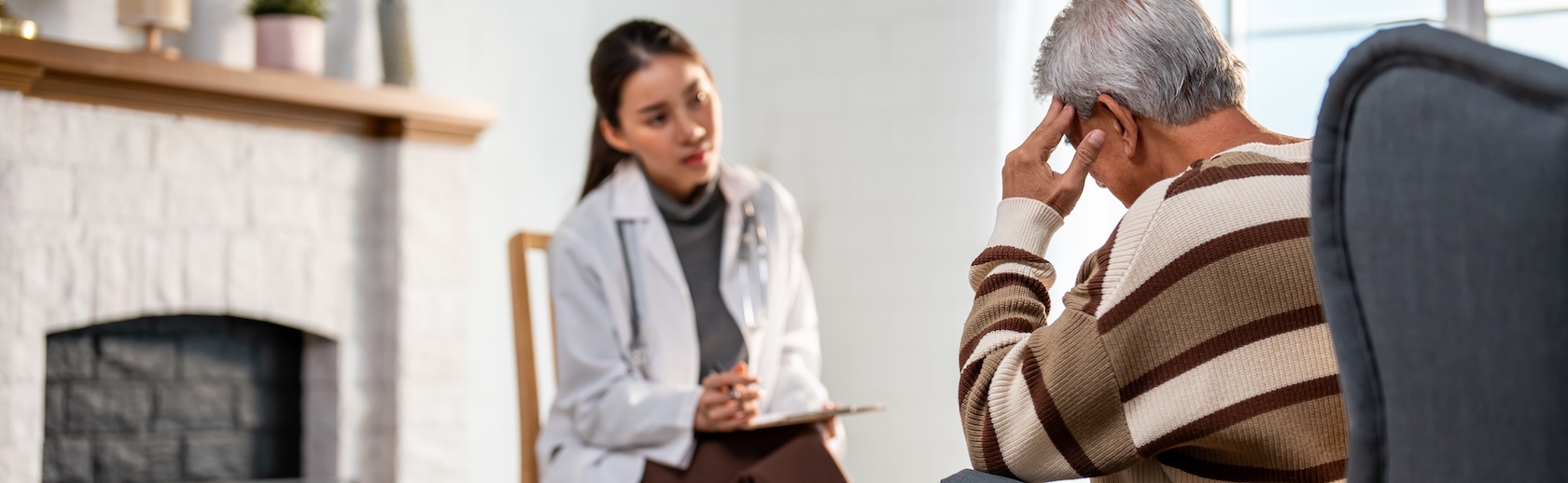 A female doctor in a white coat is consulting with a senior male patient holding his head in a brown striped sweater, offering intervention support services.