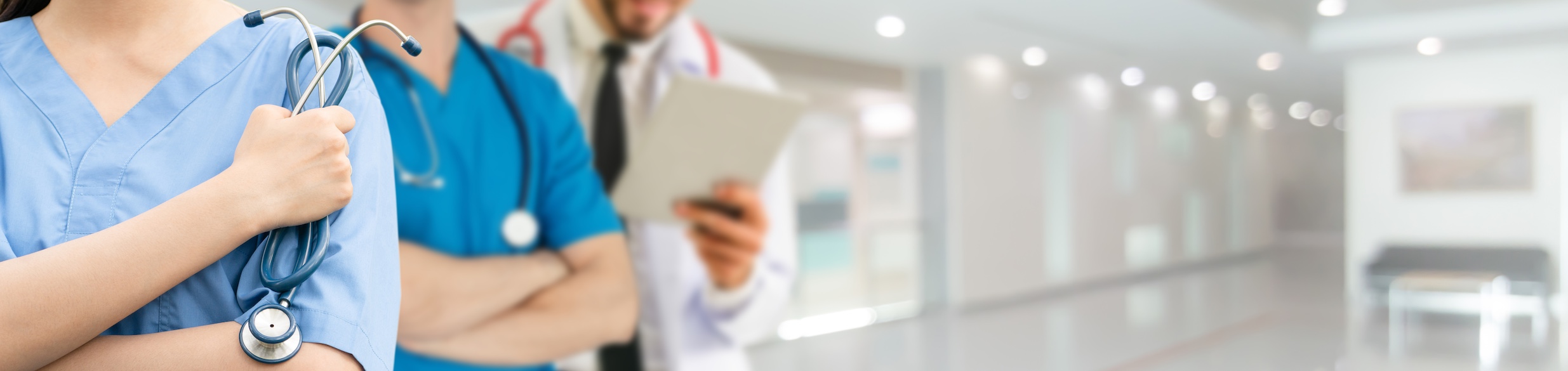 Three medical professionals in blue scrubs are holding stethoscopes, standing in a bright modern hospital corridor, representing the substance abuse treatment coverage and healthcare team.