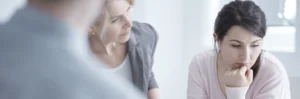 An older female counselor in a gray cardigan is comforting a thoughtful young woman in a pink sweater during a supportive therapy session for a successful journey through recovery.