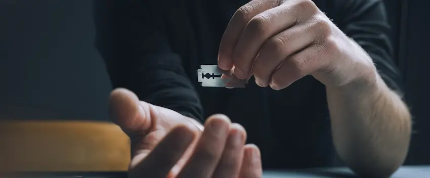 A man in a dark shirt holds a razor blade over his other hand in a dimly lit setting depicting the dangerous self-destructive behaviors that men's addiction treatment programs are designed to address and prevent at Carolina Recovery in North Carolina.