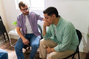 A man in a plaid shirt places a supportive hand on the shoulder of a visibly distressed man seated beside him in a group therapy circle representing the peer support and compassionate care central to effective men's addiction treatment programs at Carolina Recovery in North Carolina.